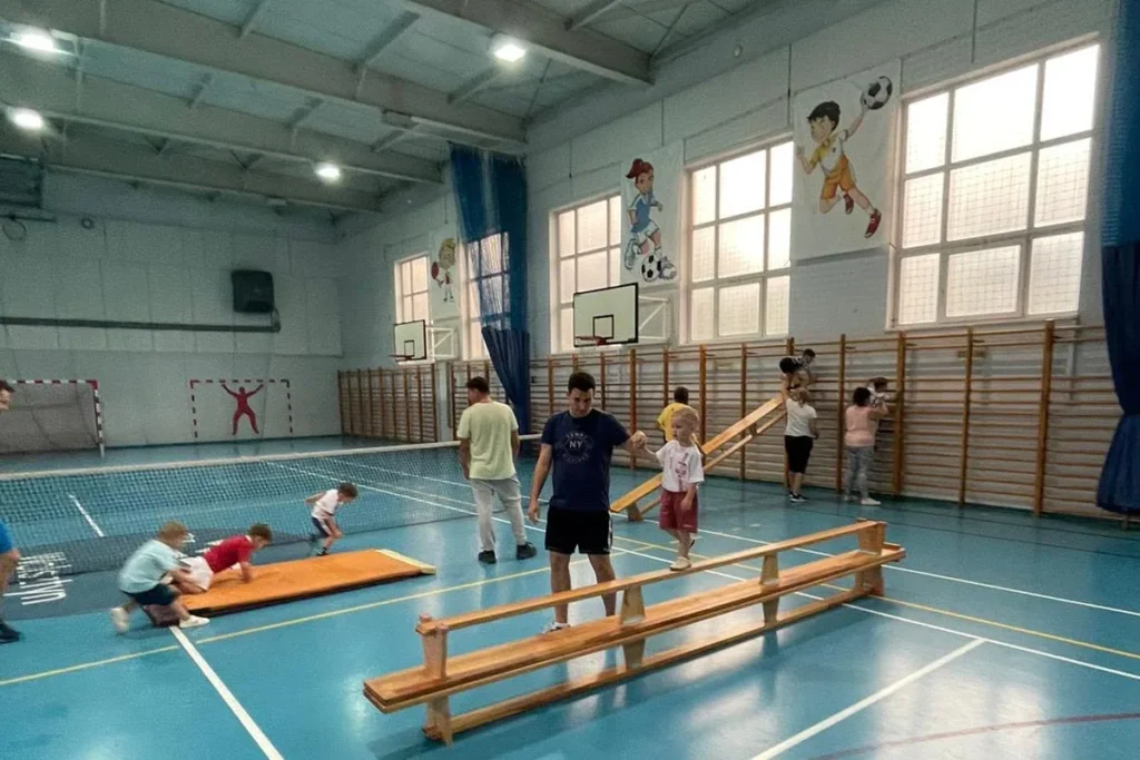 Duża sala gimnastyczna z ławeczkami i drabinkami, dzieci ćwiczą z trenerem podczas zajęć Ferajny