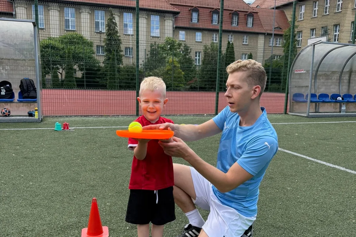Trener klęczy obok chłopca i pokazuje mu ćwiczenie z paletką i piłeczką na boisku
